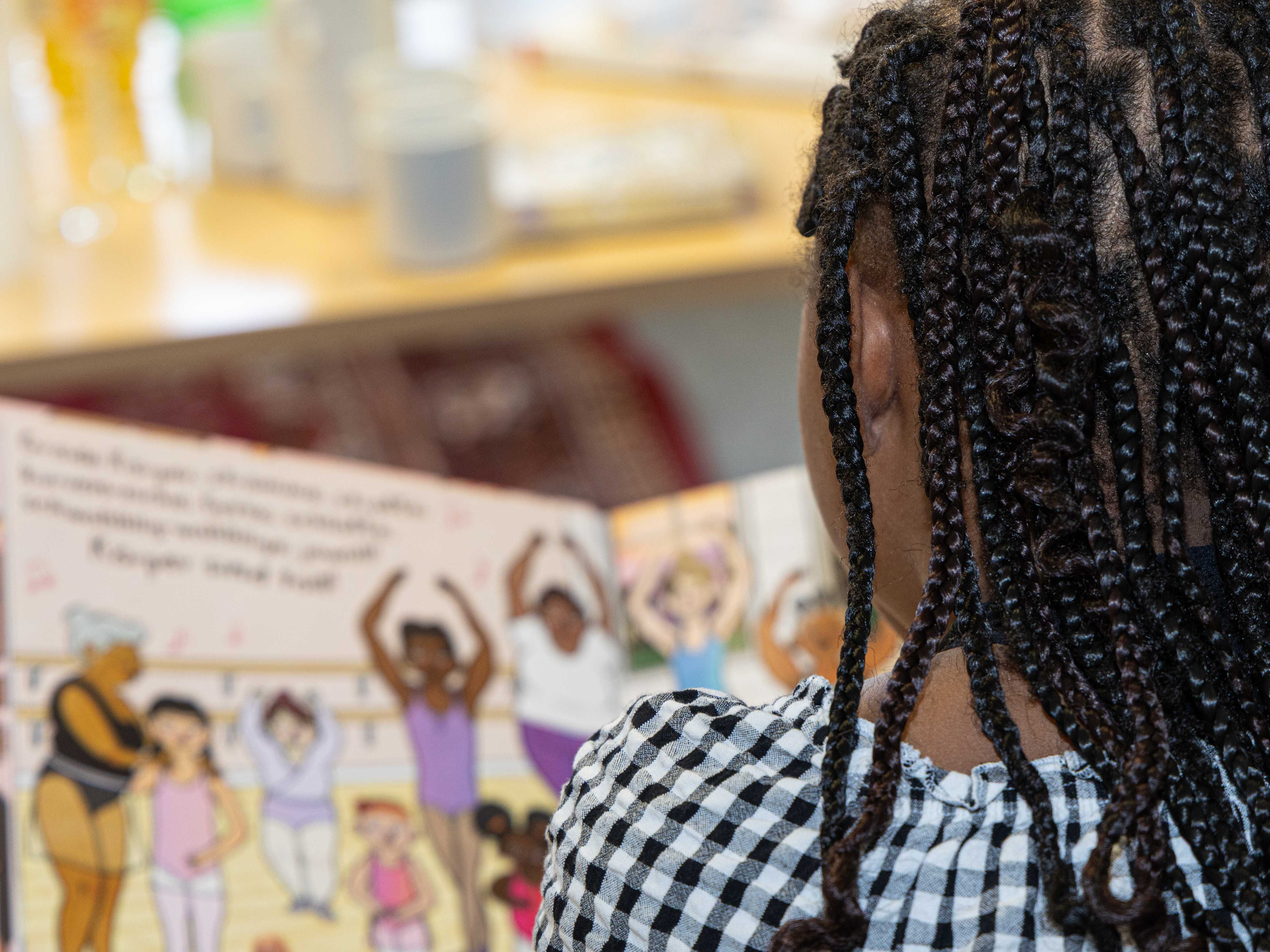 Ein Kind mit Afrohaaren und geflochtenen Zöpfen sitzt mit dem Rücken zur Kamera und betrachtet ein aufgeschlagenes Bilderbuch. Das Kind trägt ein schwarz-weiß kariertes Oberteil. Im Buch sind mehrere gezeichnete Figuren mit unterschiedlichen Hauttönen und Frisuren zu sehen, die gemeinsam in einer Reihe stehen. Im Hintergrund stehen unscharf Geschirr und Gläser auf einem Tisch.