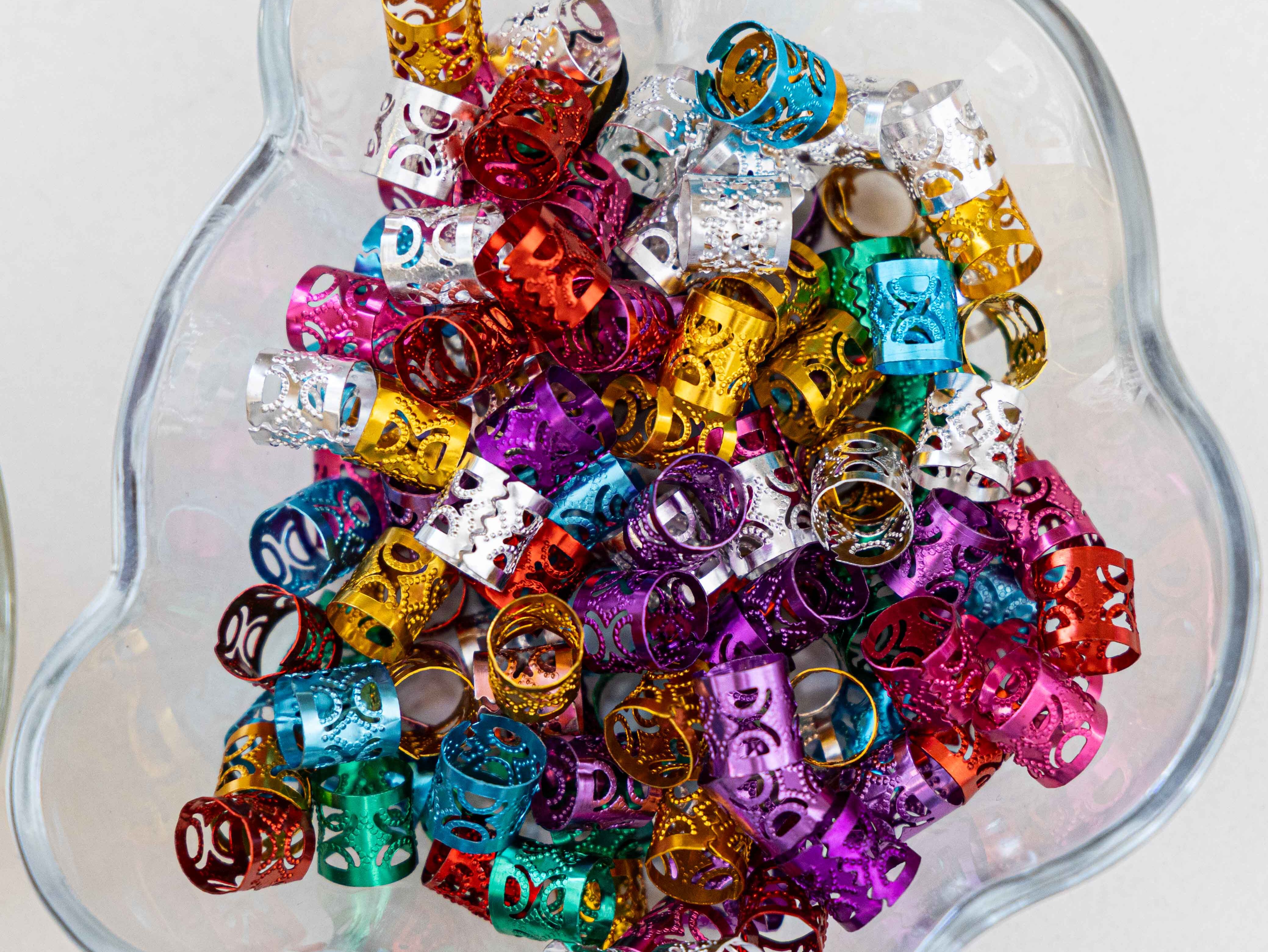 Bunte Haarklammern aus dünnem, glänzendem Metall liegen in einer transparenten, blütenförmigen Schale. Die Haarklammern sind zylinderförmig, mit ausgestanzten Mustern und in verschiedenen Farben wie Gold, Silber, Pink, Lila, Grün, Blau und Rot gehalten. Die Schale steht auf einer hellen, glatten Oberfläche.