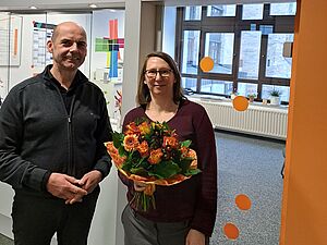 Begrüßung der neuen Leitung der kefb An der Ruhr: Zwei Personen stehen in einem modernen Büroflur. Eine Person hält einen bunten Blumenstrauß mit orangefarbenen und roten Blüten, als Zeichen der Wertschätzung und des Willkommens. Im Hintergrund sind Glaswände, farbige Dekorationselemente und ein Fenster zu sehen, die eine freundliche und offene Atmosphäre vermitteln.