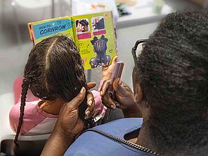 Eine erwachsene Person mit dunkler Haut und kurzen, krausen Haaren flechtet einem Kind mit langen, glatten Afrohaaren Zöpfe. Die erwachsene Person hält einen Kamm in der Hand. Das Kind hält ein farbig illustriertes Buch mit dem Titel 'How to Cornrow' aufgeschlagen, das Schritt-für-Schritt-Anleitungen und Fotos zum Flechten von Cornrows zeigt. Im Hintergrund sind unscharf weitere Personen und Möbelstücke zu erkennen.