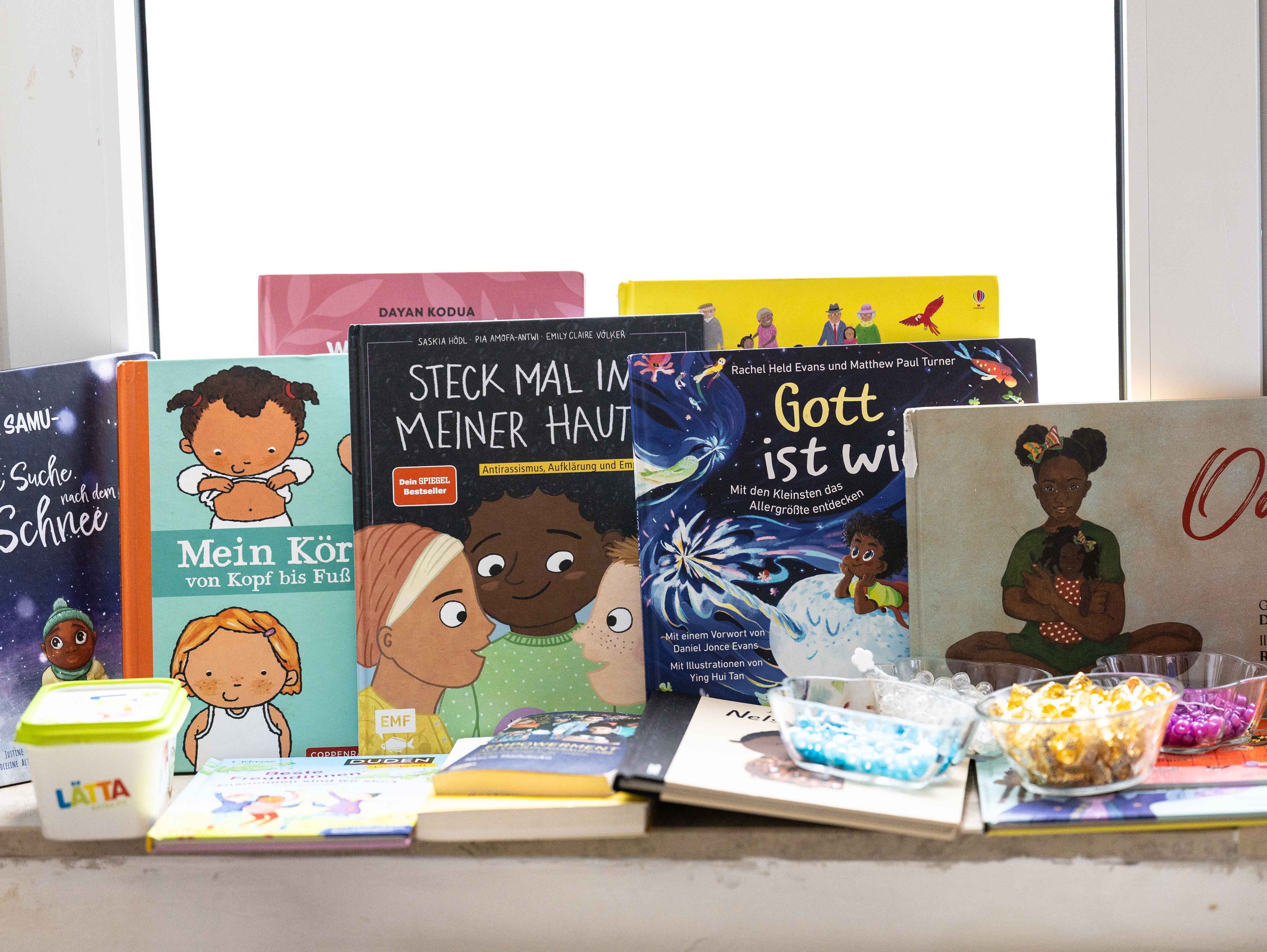 Verschiedene Kinderbücher mit Illustrationen von Kindern mit Afrohaaren stehen nebeneinander auf einer Fensterbank. Die Buchcover zeigen vielfältige Darstellungen von Kindern mit unterschiedlichen Hauttönen und Afrofrisuren. Einige Bücher sind aufrecht aufgestellt, andere liegen flach. Davor stehen kleine Schalen mit bunten Perlen und Haaraccessoires. Die Szene ist hell und freundlich, der Fokus liegt auf den Büchern und den Haaraccessoires.
