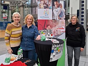 Drei Personen stehen an einem Informationsstand in einer modernen Glaspassage. Der Stand gehört zur kefb An der Ruhr und präsentiert das Projekt „Wellcome – Praktische Hilfe nach der Geburt“. Auf dem Tisch liegen Informationsmaterialien, Flyer und ein rotes T-Shirt mit dem Wellcome-Logo. Im Hintergrund ist ein großer Banner mit Fotos von Familien und Babys sowie dem Wellcome-Logo zu sehen. Die Umgebung wirkt offen und lichtdurchflutet, mit hohen Glasfassaden und einer städtischen Architektur.