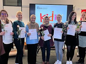 Eine Gruppe von sieben Personen steht in einem Seminarraum nebeneinander. Jede Person hält ein Zertifikat und eine Rose in der Hand. Im Hintergrund ist ein großer Bildschirm zu sehen, auf dem das Logo „kefb“ und „Kidix – Kinder. Eltern. Kurs.“ angezeigt wird. Links steht ein Flipchart, rechts eine rote Pinnwand.