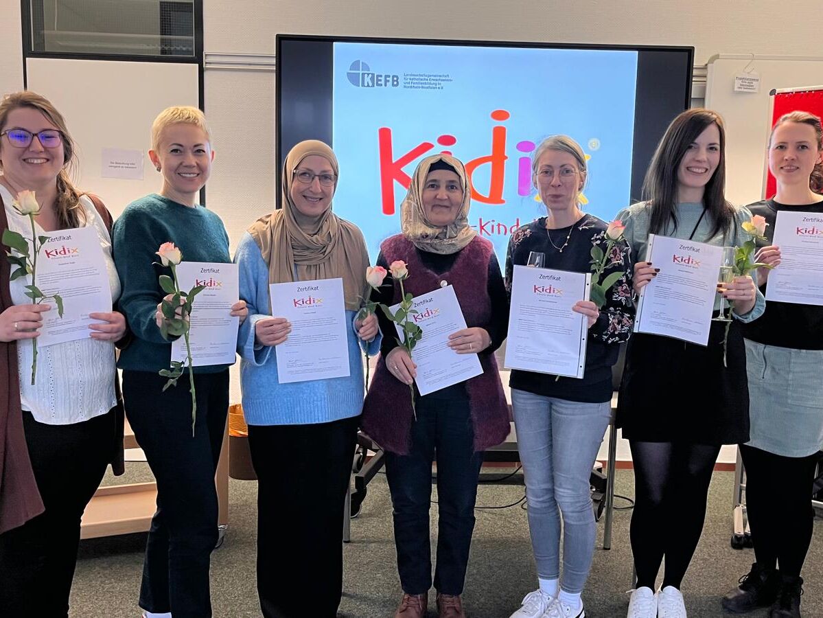 Eine Gruppe von sieben Personen steht in einem Seminarraum nebeneinander. Jede Person hält ein Zertifikat und eine Rose in der Hand. Im Hintergrund ist ein großer Bildschirm zu sehen, auf dem das Logo „kefb“ und „Kidix – Kinder. Eltern. Kurs.“ angezeigt wird. Links steht ein Flipchart, rechts eine rote Pinnwand.