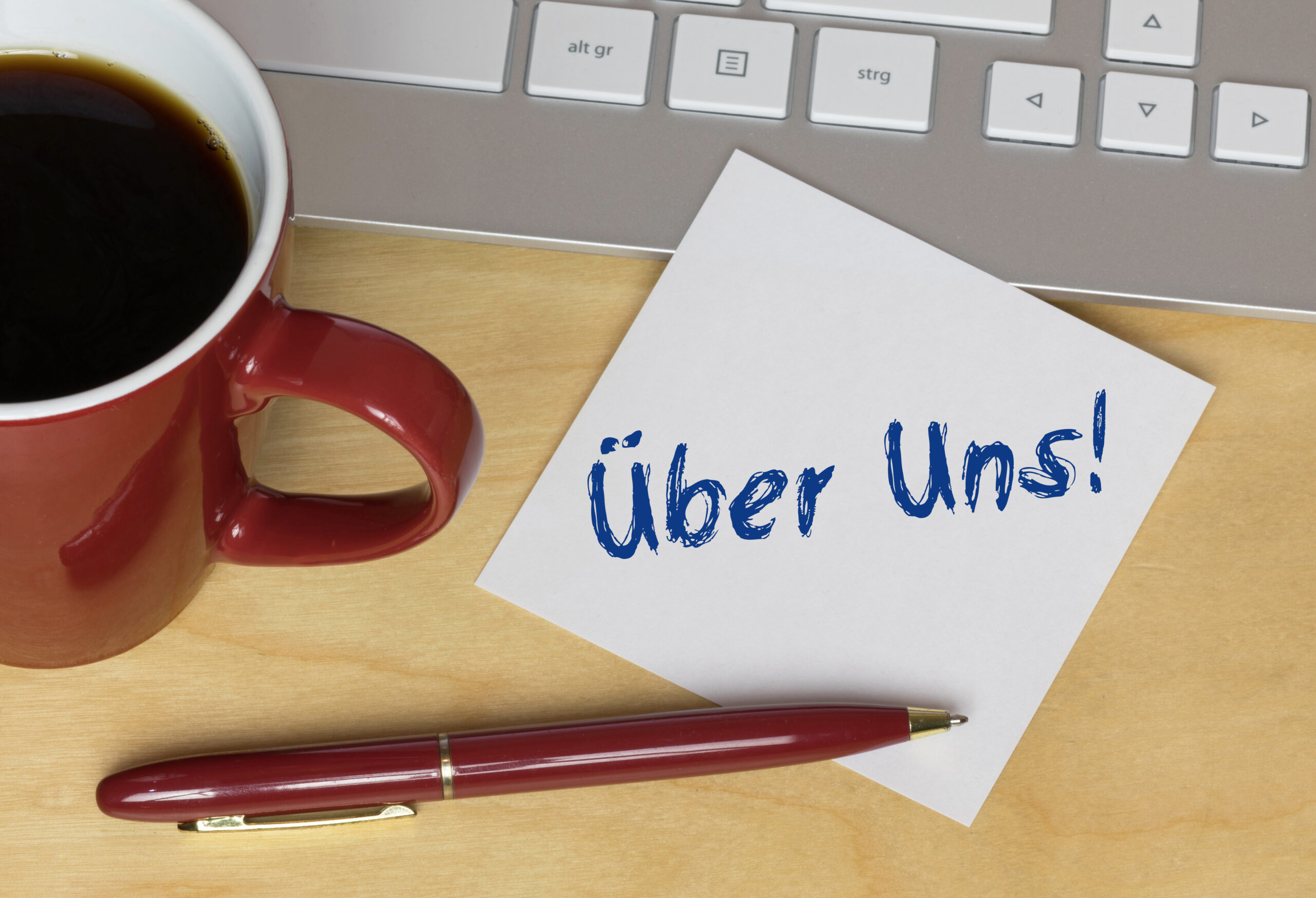 Notizzettel mit der Aufschrift ‚Über uns‘ liegt neben Kaffeetasse, Stift und Tastatur auf einem Schreibtisch.