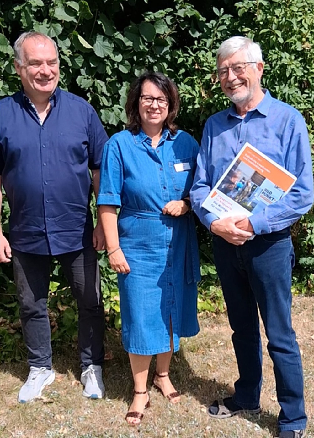 3 Personen stehen nebeneinander im Sonnenschein vor einer grünen Hecke und lächeln in die Kamera. Die Person rechts im Bild hält eine Mappe mit Unterlagen rund um die BildPunkt-Arbeit in der Hand.