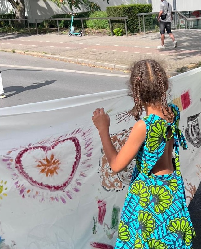 Ein Kind mit geflochtenen Haaren trägt ein ärmelloses Kleid mit blau-grünem, großflächigem Blumenmuster. Es hält ein bemaltes Banner, auf dem unter anderem ein großes Herz mit rot-gelbem Muster zu sehen ist. Die Szene spielt sich im Freien auf einer Straße ab. Im Hintergrund sind ein Gehweg, ein E-Scooter und eine weitere Person zu erkennen. Die Aufnahme entstand bei der Parade der Vielfalt in Dortmund, an der die afro-internationale Eltern-Kind-Gruppe der kefb teilgenommen hat.