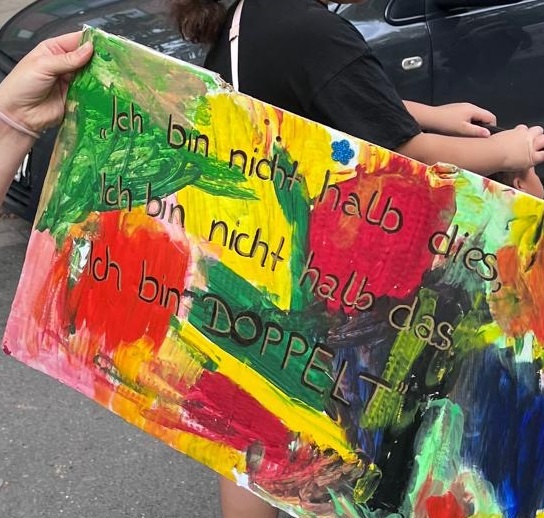 Eine Person hält ein rechteckiges, handgemaltes Schild mit bunten Farbflächen in Rot, Gelb, Grün und Blau. Auf dem Schild steht in klar lesbarer Schrift: 'Ich bin nicht halb dies Ich bin nicht halb das Ich bin DOPPELT'. Im Hintergrund sind Teile von zwei Personen und ein graues Auto zu sehen. Die Szene spielt im Rahmen der Parade der Vielfalt in Dortmund, an der die afro-internationale Eltern-Kind-Gruppe der kefb teilnimmt.