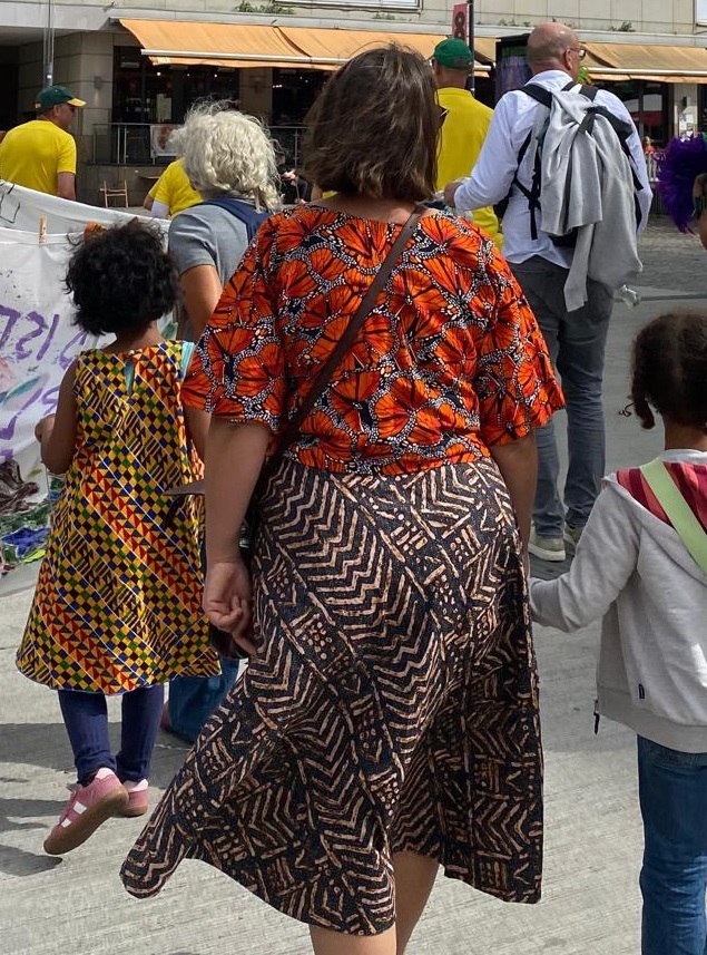 Im Vordergrund steht eine Frau mit schulterlangem Haar, die ein farbenfrohes Oberteil mit orangefarbenem, afrikanisch inspiriertem Muster und einen Rock mit geometrischem Muster trägt. Sie hält ein Kind an der Hand, das einen hellen Pullover und eine blaue Hose trägt. Links daneben läuft ein weiteres Kind mit einem Kleid in gelb-schwarzem Muster. Im Hintergrund sind mehrere Erwachsene und Kinder zu sehen, einige tragen gelbe T-Shirts. Die Szene spielt im Rahmen der Parade der Vielfalt in Dortmund, an der die afro-internationale Eltern-Kind-Gruppe der kefb teilnimmt. Die Gruppe bewegt sich gemeinsam auf einer Straße, im Hintergrund sind Gebäude und ein Banner mit bunten Buchstaben sichtbar.