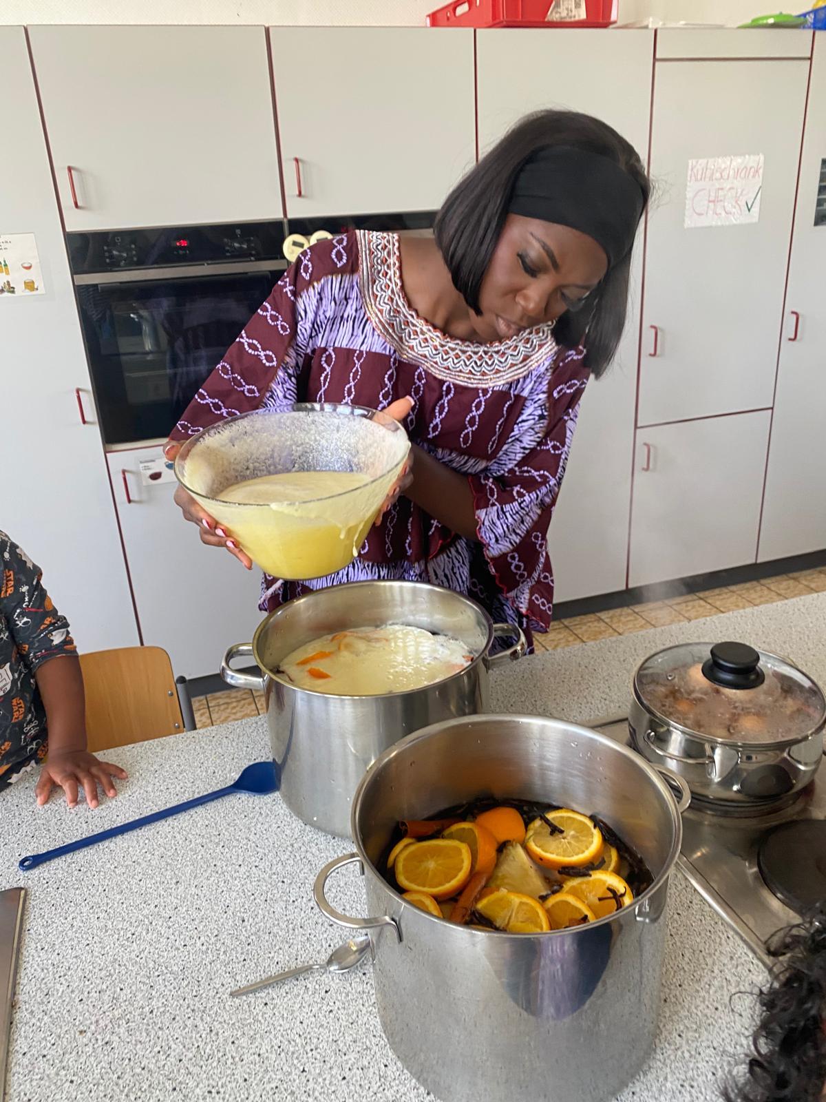 Eine Frau in traditioneller afrikanischer Kleidung gießt eine gelbe Flüssigkeit aus einer Schüssel in einen großen Kochtopf. Auf der Arbeitsfläche stehen weitere Töpfe, einer davon enthält Wasser mit Orangenscheiben und anderen Früchten. Im Hintergrund sind Küchenschränke und ein Backofen zu sehen. Die Szene zeigt einen afrikanischen Koch-Workshop.