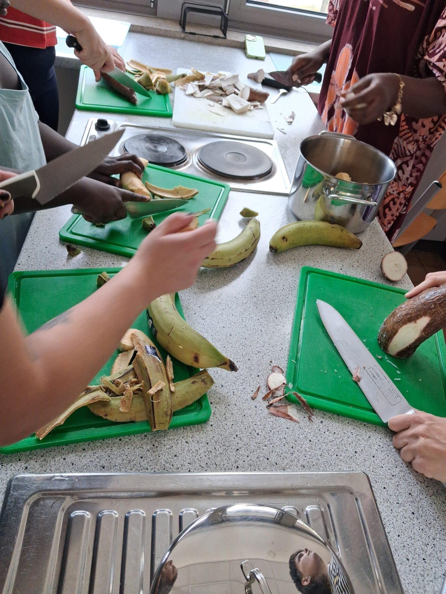 Im Mittelpunkt steht eine Küchenszene während eines afrikanischen Koch-Workshops. Mehrere Personen schneiden mit großen Messern Kochbananen und Yamswurzeln auf grünen Schneidebrettern. Die Zutaten werden geschält und zerkleinert, einige Stücke liegen bereits auf dem Tisch. Im Hintergrund sind ein Edelstahltopf, ein Herd und weitere Küchenutensilien zu sehen. Die Kleidung der Teilnehmenden ist unterschiedlich, darunter ein gemustertes Oberteil.