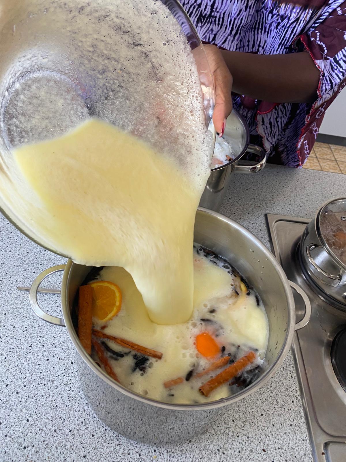 Eine Person gießt eine cremig-gelbe Flüssigkeit aus einer großen Schüssel in einen Kochtopf. Im Topf schwimmen mehrere Zimtstangen, eine Orangenscheibe und weitere Gewürze in einer hellen, schaumigen Flüssigkeit. Die Szene spielt auf einer Küchenarbeitsplatte neben einem Herd.