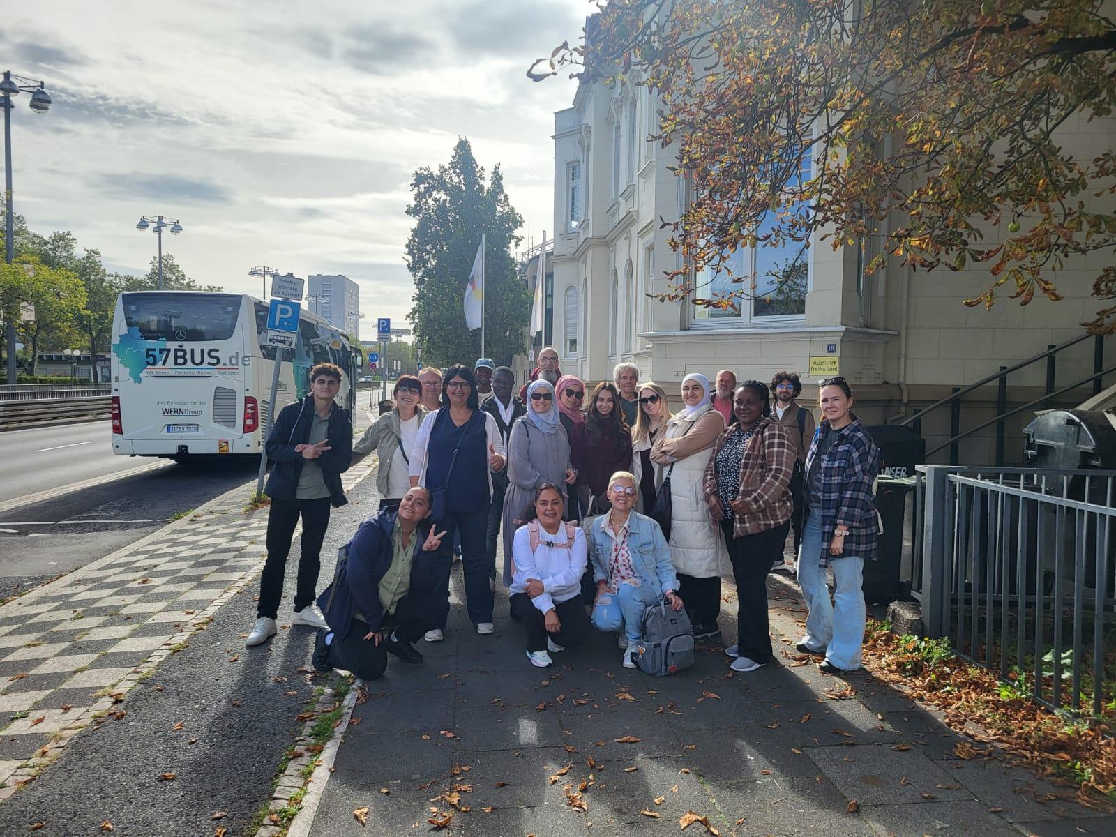 Eine Gruppe von Menschen steht auf einem Gehweg vor einem hellen Gebäude mit klassizistischer Fassade. Im Hintergrund ist ein weißer Reisebus mit der Aufschrift '57BUS.de' zu sehen, der am Straßenrand parkt. Die Personen sind unterschiedlich gekleidet, einige tragen Jacken oder Sonnenbrillen, andere halten Taschen oder Rucksäcke. Herbstlich gefärbte Blätter hängen an einem Baum rechts im Bild. Die Szene spielt tagsüber bei bewölktem Himmel. Die Gruppe besteht aus Teilnehmenden des Projekts 'Labor Ankommen' während eines Bildungsausflugs in Bonn.