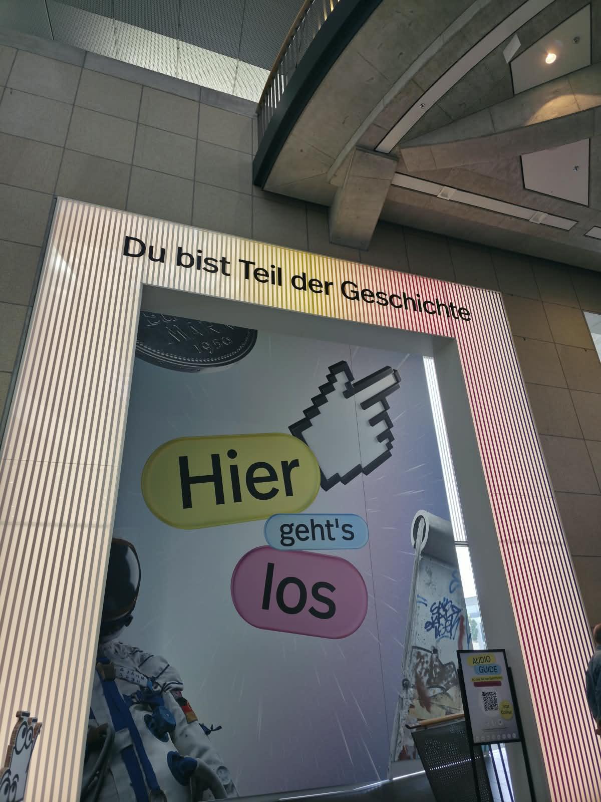 Ein großer, rechteckiger Durchgang mit leuchtenden, vertikal verlaufenden Lichtstreifen rahmt den Eingangsbereich. Über dem Durchgang steht in klaren Buchstaben: 'Du bist Teil der Geschichte'. Dahinter ist eine Wand mit einer bunten Grafik zu sehen, auf der eine große, weiße Hand als Cursor auf eine Sprechblase mit der Aufschrift 'Hier geht's los' zeigt. Im unteren Bereich ist ein Teil eines Raumanzugs zu erkennen. Die Architektur im Hintergrund wirkt modern und besteht aus Beton und Glas. Das Bild entstand im Rahmen eines Bildungsausflugs der Teilnehmenden des Projekts 'Labor Ankommen' nach Bonn.