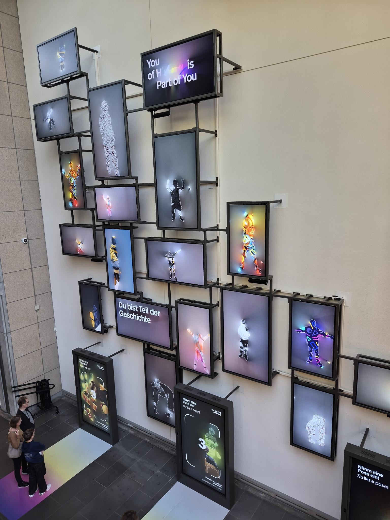 Mehrere beleuchtete Bildschirme sind an einer Wand in einem öffentlichen Gebäude angebracht. Auf den Monitoren sind digital bearbeitete Porträts von Menschen zu sehen, die in unterschiedlichen Farben und Mustern gestaltet sind. Zwei Personen stehen unten links und betrachten die Installation. Auf einem der Bildschirme steht der Text: 'Du bist Teil der Geschichte'. Die Szene zeigt eine moderne Ausstellung, die Teil des Bildungsausflugs der Teilnehmenden des Projekts 'Labor Ankommen' nach Bonn ist.